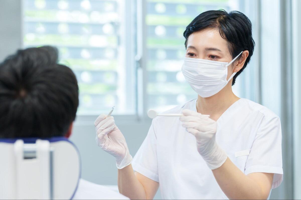 歯科医院にて、マスクと手袋を着用した歯科衛生士が器具を手に持ち、診察台の患者へ説明をしている様子