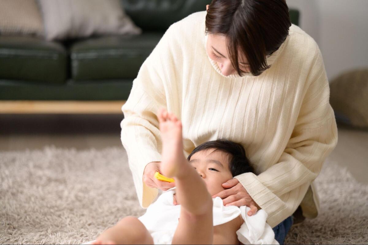 リビングのカーペットの上で、仰向けに寝ている赤ちゃんの口元に黄色い歯ブラシをあてて仕上げ磨きをする母親