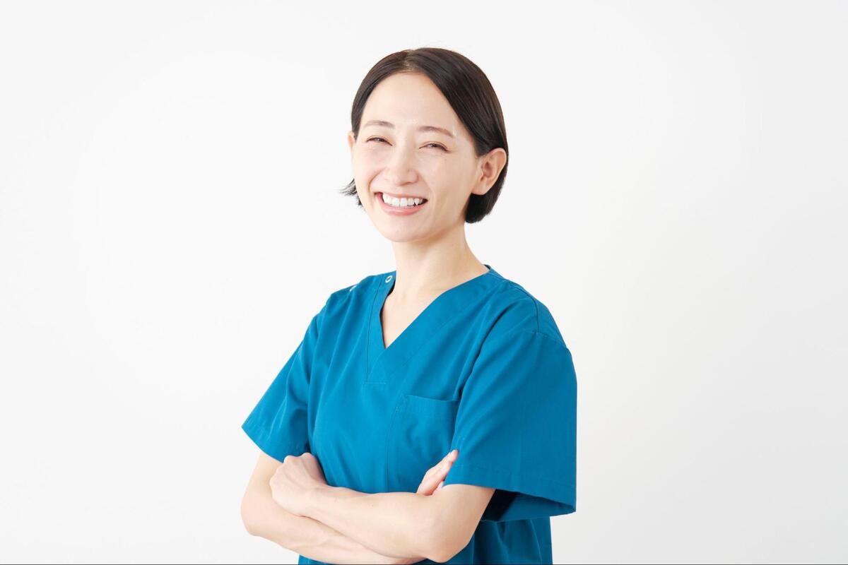 白背景の前で、青いスクラブを着用した歯科衛生士が腕を組み、カメラに向かって微笑んでいる様子