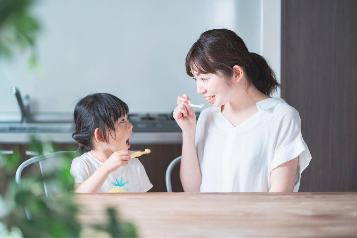 食卓でお母さんと向かい合い、お互いに歯ブラシを持って笑顔で楽しく歯磨きの練習をする親子の様子