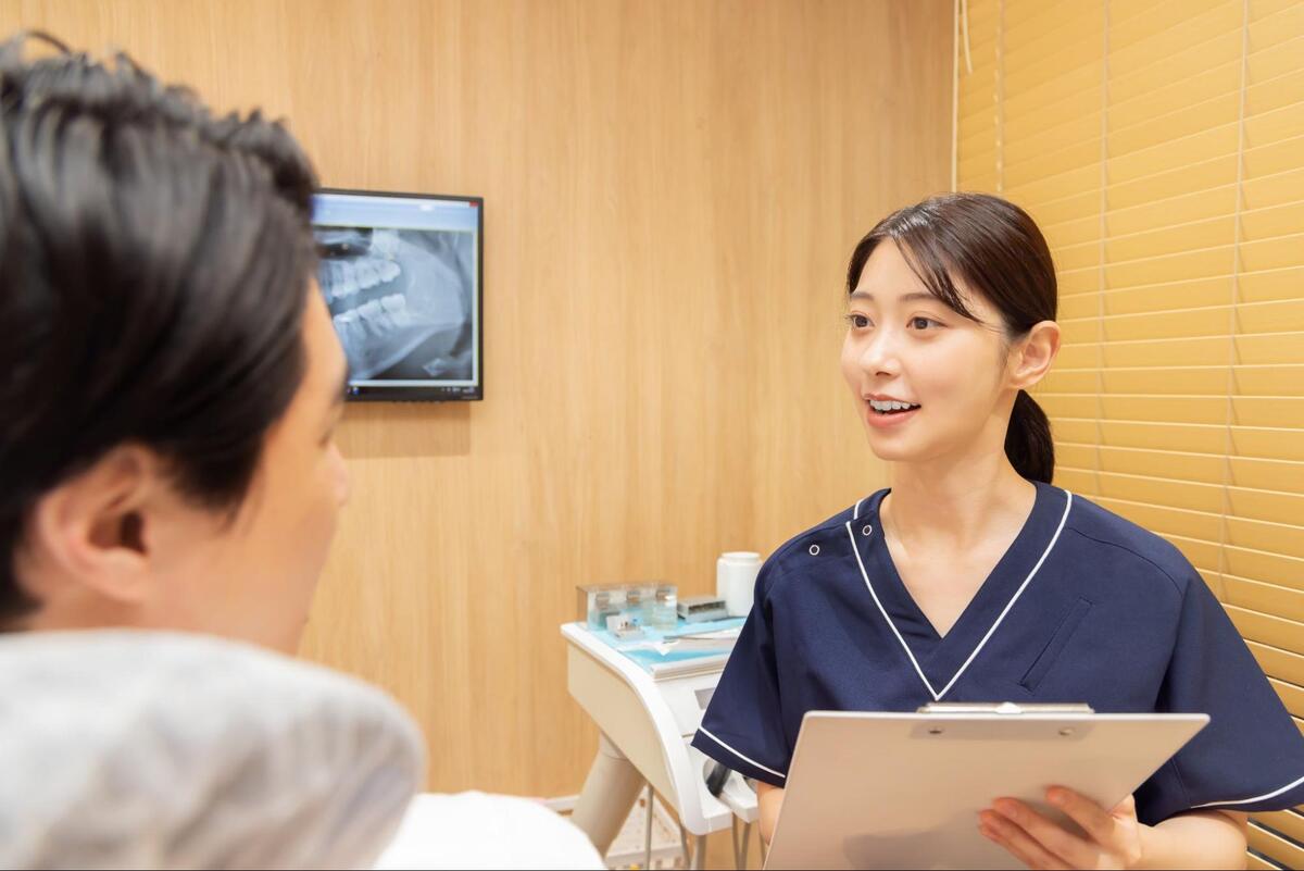 歯科診療室にて、歯科衛生士がカルテを手に持ちながら患者に治療の説明をしている様子