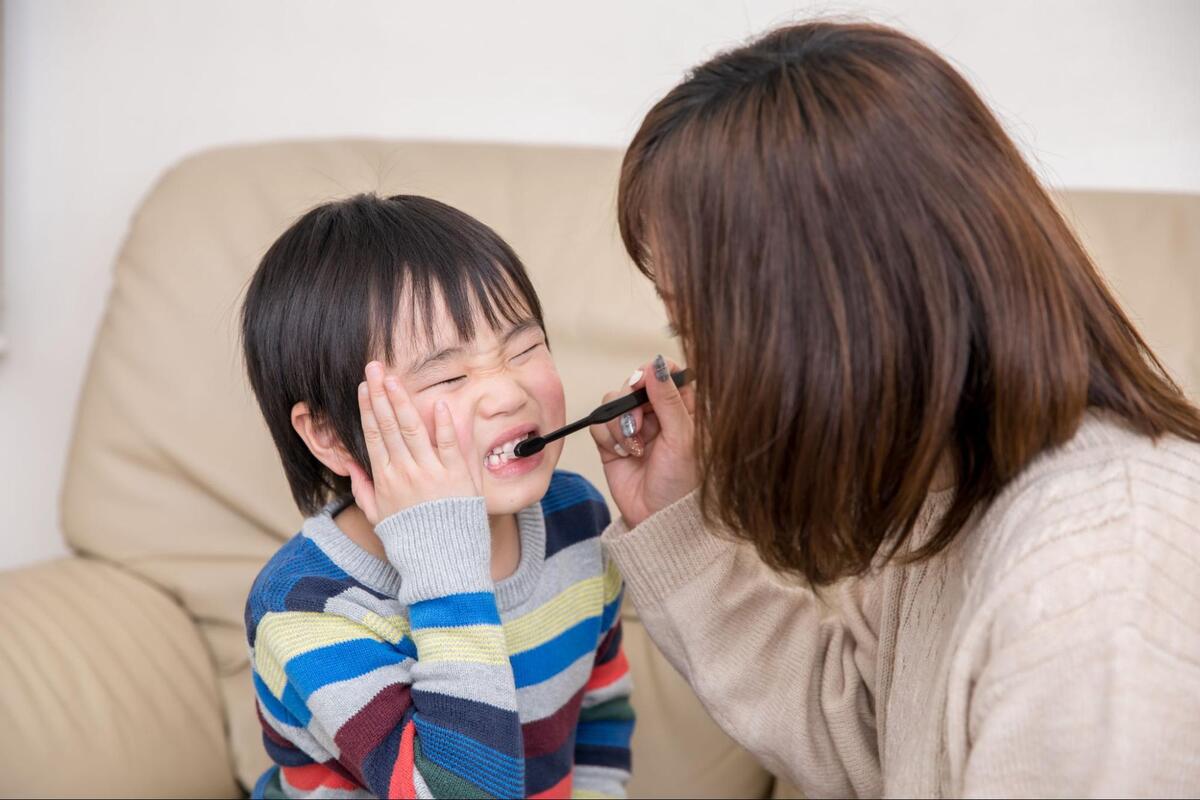 ソファの前で頬に手を当てて口を開ける男の子と、黒い歯ブラシを使って仕上げ磨きをしてあげる母親