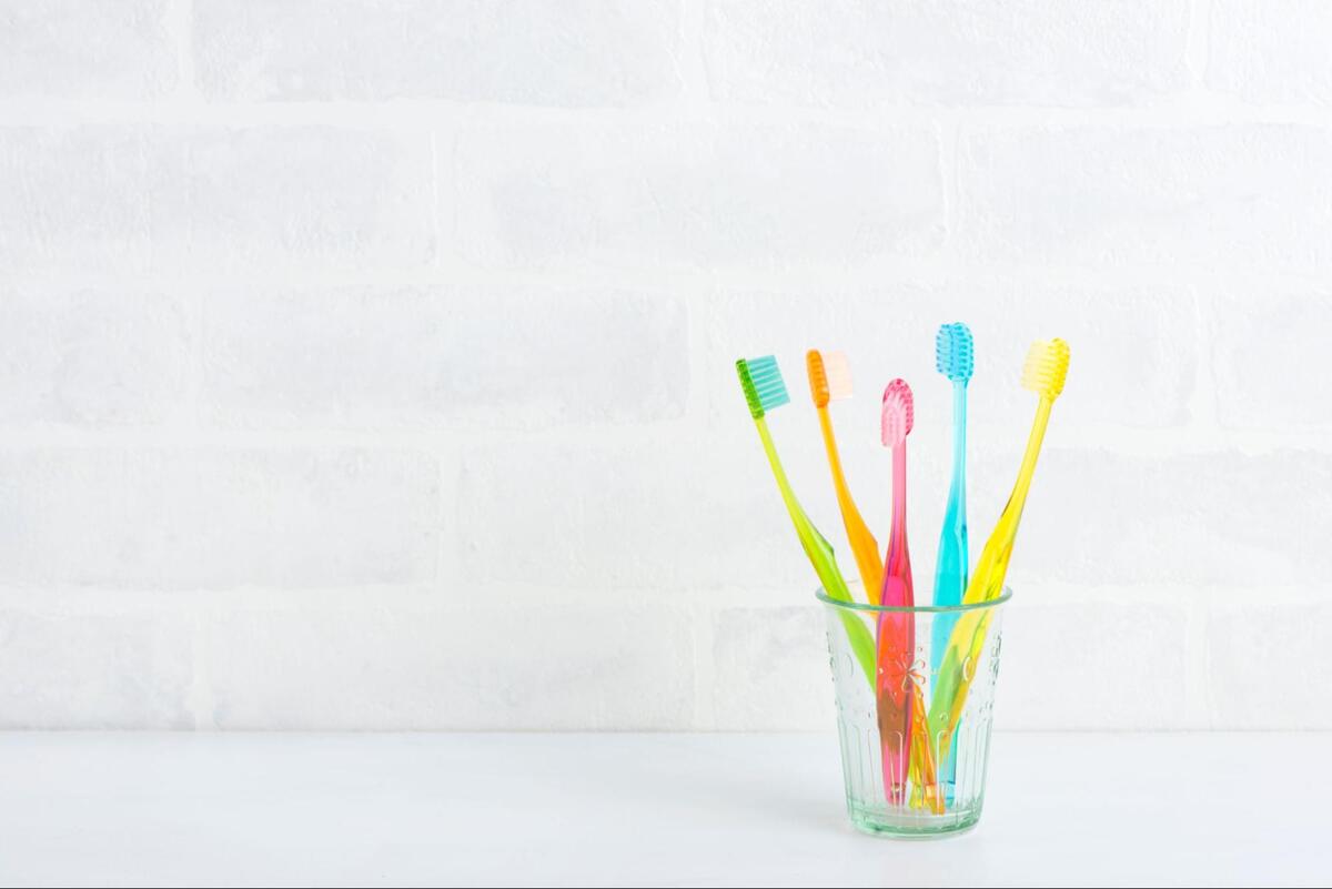 白いレンガ調の壁を背景に、透明なガラスコップに立てられたカラフルな5本のプラスチック製歯ブラシ