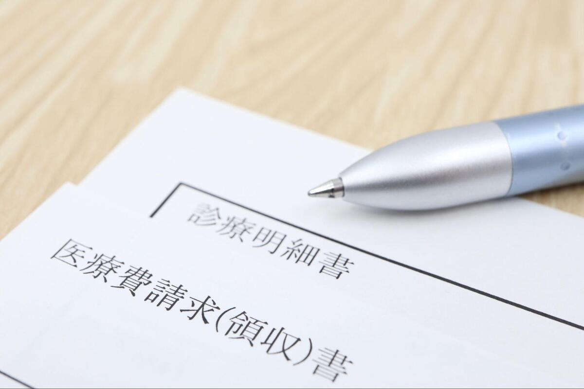 木目調のテーブルの上に置かれた「診療明細書」「医療費請求(領収)書」の文字が書かれた書類とボールペンの先端