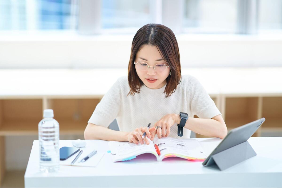 白いデスクにて、メガネをかけた女性がタブレットやスマートフォンを傍らに置き、開いたテキストを熱心に読み込んでいる様子