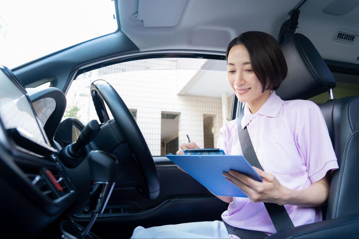 車内で、シートベルトを着用した制服姿の歯科衛生士が青いバインダーの書類にペンで記入している様子